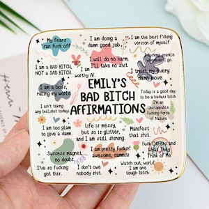 Könnte beinhalten: Ein quadratisches Keramik-Schmucktablett mit goldenem Rand, mit dem Text "Emily's Bad Bitch Affirmations" in der Mitte. Das Tablett ist mit verschiedenen Affirmationen sowie Blumen- und Sterndesigns verziert. Das Tablett wird von einer Person gehalten.