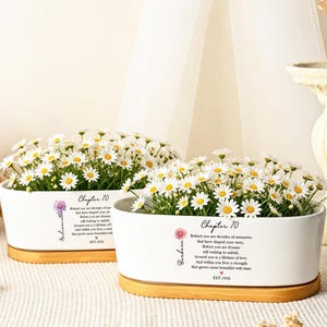 May include: Two oval white ceramic planters with bamboo bases, each filled with white daisy flowers. The planters feature the text 'Chapter 70', a floral graphic, a personalized name, and the text 'Behind you are decades of memories' with 'EST. 1956'.