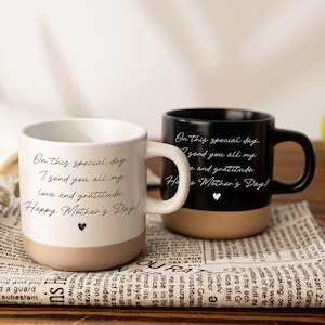 Puede incluir: Dos tazas de cerámica con base beige y asa blanca. Una taza es blanca con texto negro que dice: "En este día especial, te envío todo mi amor y gratitud. ¡Feliz Día de la Madre!" La otra taza es negra con el mismo texto.