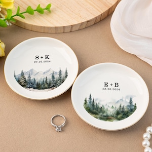 Peut inclure: Deux coupelles à anneaux en céramique blanche avec une bordure dorée. Chaque coupelle présente une illustration aquarelle d'une forêt avec des montagnes en arrière-plan. La coupelle du haut porte le texte "S+K 07.16.2024" et la coupelle du bas porte le texte "E+B 05.25.2024".