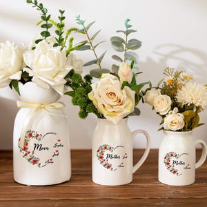 Puede incluir: Tres jarrones de cerámica blanca de diferentes tamaños, cada uno con flores. Los jarrones tienen un diseño floral en forma de corazón con texto. El jarrón más grande tiene una cinta. Las flores incluyen rosas blancas y crema.