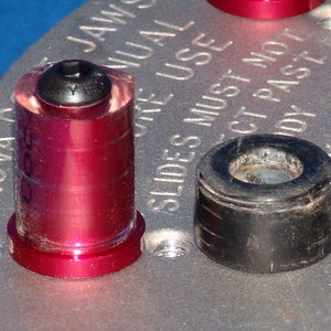 May include: A close-up of two metal and plastic components. One is a red cylinder with a black top, and the other is a black cylindrical piece. The components are on a silver surface with engraved text.