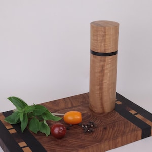 May include: A cylindrical wooden pepper mill with a light brown grain and a thin black horizontal band. It sits on a rectangular wooden cutting board featuring a dark brown wood grain, black side panels, and small light wood square inlays.