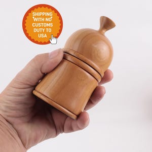 Puede incluir: Un molinillo de pimienta de madera marrón claro con un cuerpo bulboso y una parte superior pequeña y redondeada. El molinillo se sostiene en una mano sobre un fondo blanco. El texto "SHIPPING WITH NO CUSTOMS DUTY TO USA" está en un gráfico circular naranja.