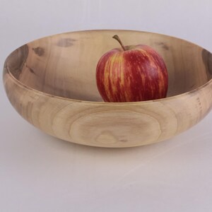 Könnte beinhalten: Eine handgefertigte Holzschale mit natürlichem Finish, in der sich ein rot-gelber Apfel befindet. Die Schale hat eine glatte, abgerundete Form und sichtbare Holzmaserung. Der Apfel ist reif und hat einen Stiel.