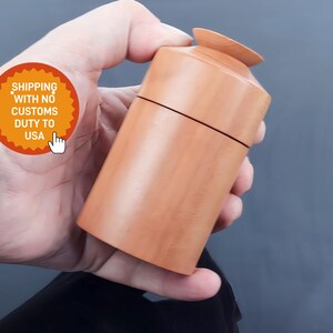 Puede incluir: Un recipiente de madera marrón claro con un cuerpo cilíndrico y una tapa pequeña y redonda. El recipiente se sostiene en una mano sobre un fondo gris. El recipiente tiene un acabado liso y una veta de madera natural.
