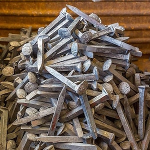 May include: A large pile of rusty, metal nails in a wooden crate. The nails are all different sizes and shapes, and some have a square head.