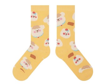 May include: A pair of yellow socks with a repeating pattern of cartoon sheep and cookies. The sheep have white fur, brown hair, and red accents. The socks are made of a soft material.