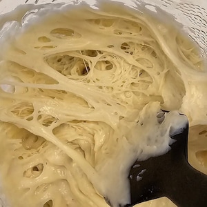 May include: A close-up of a white, stretchy dough in a glass bowl. The dough is very elastic and has a lot of air bubbles. A black spatula is in the bowl, partially submerged in the dough.