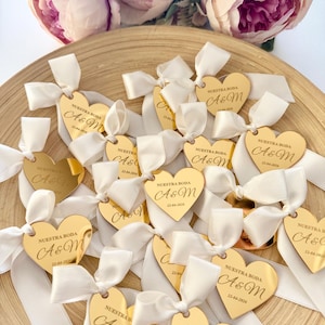 Könnte beinhalten: Goldene herzförmige Hochzeitsgeschenke mit weißen Schleifen. Jedes Herz trägt die Gravur "NUESTRA BODA A&M 22-04-2026". Die Gastgeschenke sind auf einem Holztablett angeordnet, mit rosa Blumen im Hintergrund.