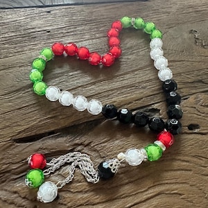 May include: A beaded necklace with red, green, white, and black beads. The beads are strung together with a silver chain and have a decorative tassel at the end. The necklace is displayed on a wooden surface.