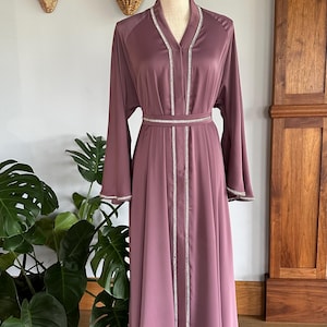 May include: A long, mauve-colored satin dress with a V-neckline and a silver beaded trim. The dress has a belt at the waist and long sleeves.