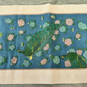 May include: A blue fabric panel with embroidered peacocks and pink flowers. The peacocks are green, blue, and gold. The flowers are pink and white.