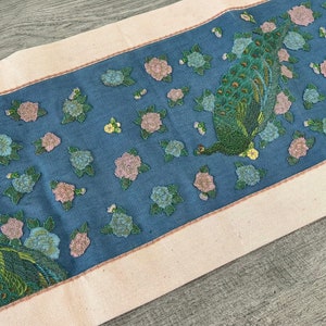 May include: A blue and white table runner with a repeating pattern of peacocks and flowers. The runner has a cream-coloured border.