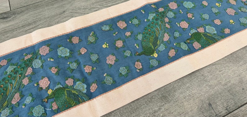 May include: A blue and white table runner with a repeating pattern of peacocks and flowers. The runner has a cream-coloured border.