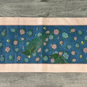 May include: A blue and pink floral embroidered table runner with a pink border. The runner features a repeating pattern of flowers and leaves.