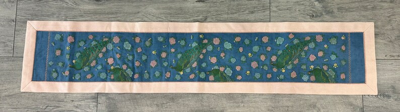 May include: A blue and pink floral embroidered table runner with a pink border. The runner features a repeating pattern of flowers and leaves.