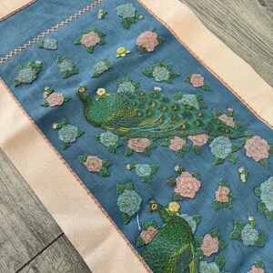May include: A blue fabric runner with a white border features an embroidered design of two peacocks with green and gold feathers surrounded by pink and blue flowers.