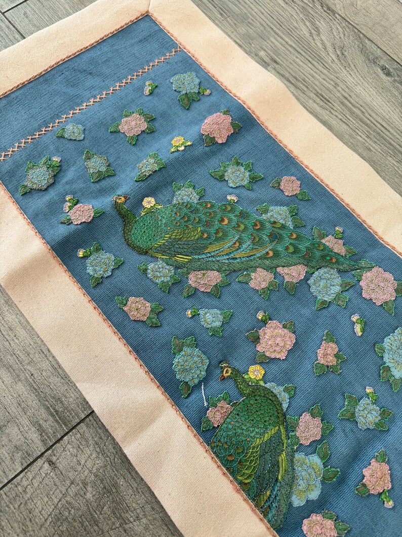 May include: A blue fabric runner with a white border features an embroidered design of two peacocks with green and gold feathers surrounded by pink and blue flowers.
