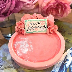 May include: A pink ceramic dish with a raised rim, adorned with sculpted pink roses and a small rectangular sign. The sign has a drawing of a smiling face and Japanese text. The dish is surrounded by pink roses.