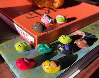 Può includere: Una collezione di piccole statuette di animali dipinte a mano in vari colori, tra cui rosso, giallo, verde, marrone, rosa e nero. Ogni statuetta presenta semplici dettagli del viso come occhi, nasi e orecchie, disposte sopra due libri.
