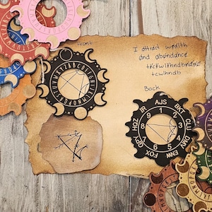 May include: A collection of circular wooden divination tools featuring moon phases and alphabet or numerical sequences in various colors including blue, red, pink, black, and green. A tan paper sheet displays the text I attract wealth and abundance.