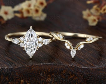 Puede incluir: Dos anillos de boda de oro. El anillo de compromiso presenta una piedra central de diamante con corte marquesa, rodeada de diamantes más pequeños. La alianza tiene un diseño curvo con pequeños detalles de diamantes. Los anillos están sobre una superficie de madera oscura.