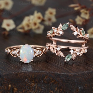 May include: Rose gold engagement ring with an oval opal center stone and diamond accents. A matching ring guard with leaf and gemstone details is also shown. The rings are displayed on a dark wooden surface.