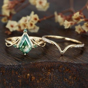 Könnte beinhalten: Ein goldfarbenes Verlobungsring-Set. Der Hauptring hat einen geometrischen, smaragdgeschliffenen grünen Stein, ergänzt durch kleinere grüne Steine und florale Details. Ein zartes, V-förmiges Band, besetzt mit kleinen, klaren Steinen, vervollständigt das Set.