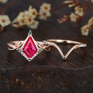 May include: A rose gold engagement ring set. The main ring features a kite-shaped, vibrant red gemstone, accented with small white diamonds and green gemstones. A matching curved band with small white diamonds completes the set. The rings are set against a dark wooden background.