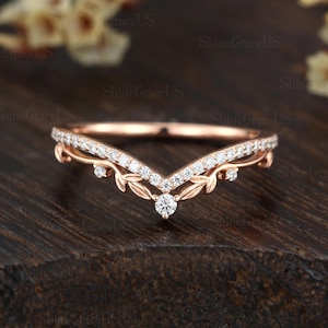 May include: A rose gold wedding ring with a V-shaped design. The ring features a row of small, sparkling diamonds along the top and a larger diamond at the center. Delicate leaf and vine details add to the ring's elegant style.