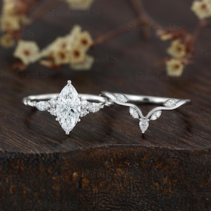 May include: A silver-toned engagement ring set featuring a central marquise-cut stone flanked by smaller round and marquise stones on a twisted band, paired with a matching V-shaped contoured wedding band adorned with tiny marquise-cut stones.