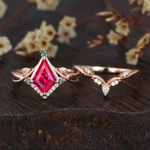 May include: A rose gold engagement ring set featuring a kite-shaped, ruby-colored center stone with a diamond halo. The set includes a matching band with a curved design and small diamonds. The rings are set on a dark wooden surface.