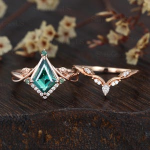 May include: Rose gold engagement ring set. The main ring features a large emerald-cut green gemstone with a diamond halo. The band has leaf accents. The matching band is curved and set with small diamonds. The rings are displayed on a dark wooden surface.