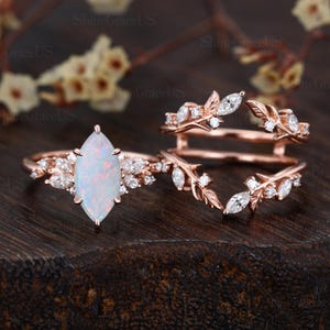 May include: A rose gold ring set featuring a marquise-cut opal center stone and accent diamonds. The set includes a main ring and a curved band, both with leaf and diamond details. The rings are displayed on a dark wooden surface.