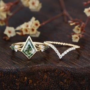 May include: Two gold rings. One ring features a geometric, green stone with a gold frame and small accent stones. The second ring is a gold band with small, white stones in a V-shape. The rings are displayed on a dark wooden surface.
