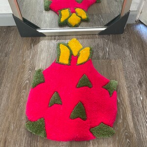 May include: A pink and green bath mat shaped like a dragon fruit. The mat has a yellow crown on top.