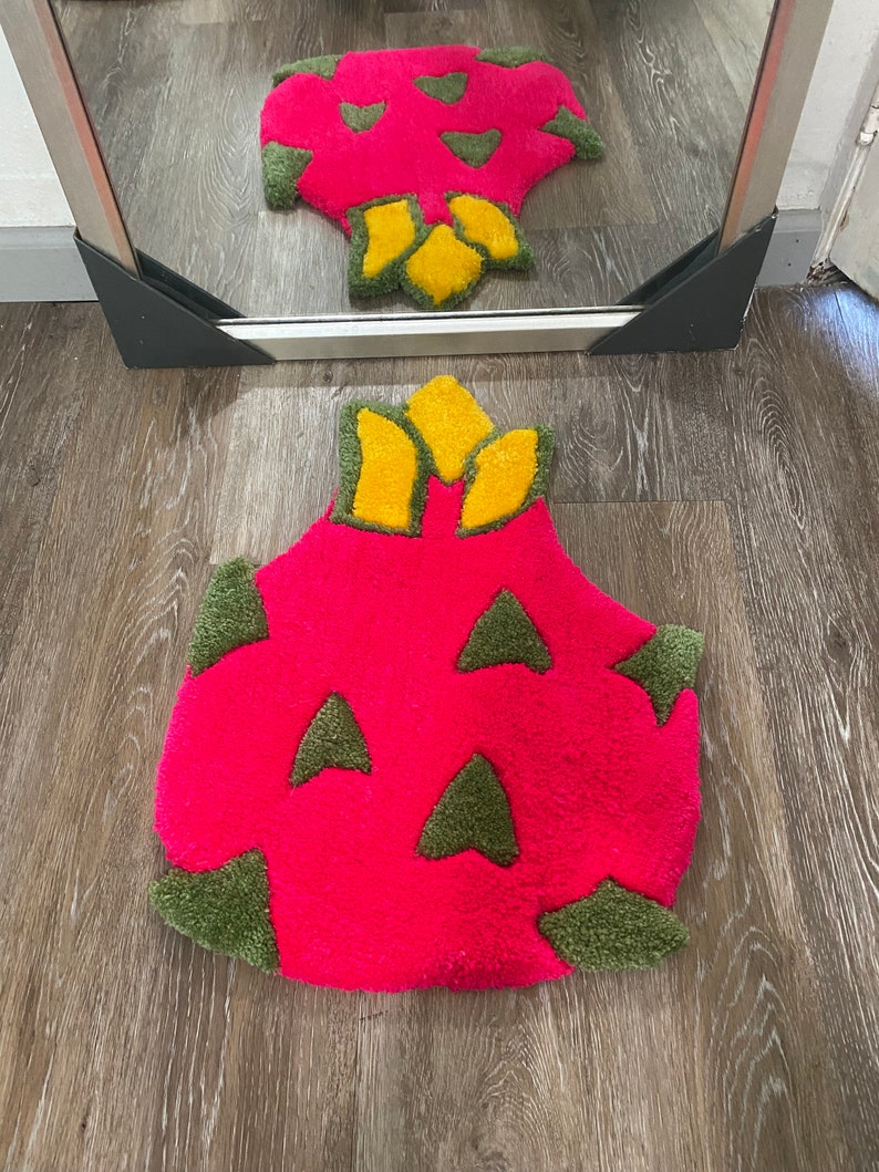 May include: A pink and green bath mat shaped like a dragon fruit. The mat has a yellow crown on top.
