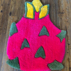 May include: A pink and green bath mat shaped like a dragon fruit. The fruit has a yellow crown on top.