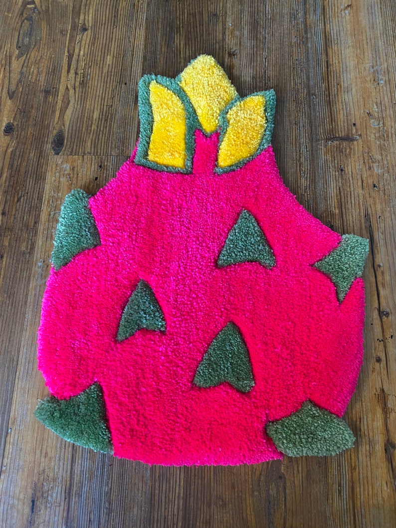 May include: A pink and green bath mat shaped like a dragon fruit. The fruit has a yellow crown on top.