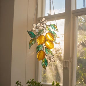 May include: Stained glass lemon branch with yellow lemons, green leaves, and white flowers. The decorative piece hangs from a silver chain in front of a window, creating a sunny, vibrant display. The glass art is a unique home decor item.
