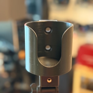 May include: A black, cylindrical holder with two circular holes, mounted on a black stand. The holder has a curved opening and a small, adjustable knob. The background is blurred, suggesting an indoor setting.