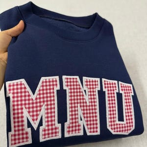 Puede incluir: Sudadera azul marino con las letras "MNU" aplicadas en la parte delantera. Las letras son de tela de cuadros rojos y blancos con un borde blanco. La sudadera tiene cuello redondo y mangas largas.