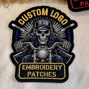 May include: A black embroidered patch featuring a skull wearing a cap and leather jacket, riding a motorcycle. The text CUSTOM LOGO is arched at the top, and EMBROIDERY PATCHES is in a rectangular box at the bottom, all in gold thread on black.