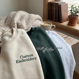 Könnte beinhalten: Drei Sweatshirts mit Rundhalsausschnitt in Creme, Dunkelgrün und Grau. Das cremefarbene Sweatshirt hat "Custom Embroidery" in Grün gestickt. Das dunkelgrüne Sweatshirt hat "Full of Love" in Weiß gestickt. Das graue Sweatshirt hat "ated" in Weiß gestickt.