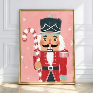 May include: A framed print of a nutcracker soldier with a candy cane. The illustration features a red uniform, black hat, and a large mustache against a pink background. White snowflakes are scattered around the figure.