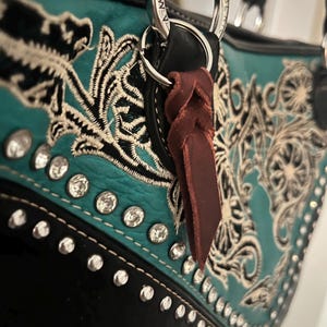 May include: A turquoise and black handbag featuring intricate white floral embroidery and a row of clear rhinestones. The bag includes a silver-toned metal ring attachment with a dark brown leather tassel hanging from the handle base.