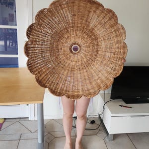 May include: A large, round, woven wicker lampshade with a scalloped edge. The lampshade is a natural tan colour and has a circular design in the centre. The lampshade is being held up by a person.
