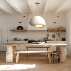 May include: A rustic wooden dining table and bench set with a large, white, round, felt pendant light hanging above. The table and bench are made of natural wood with a light brown finish. The pendant light is suspended from the ceiling by a thin cord.