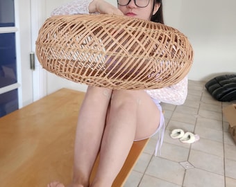 May include: An oval-shaped wicker basket. The basket is made of light brown wicker and has a lattice design. The basket is being held by a person. The person is wearing glasses and a white lace top.